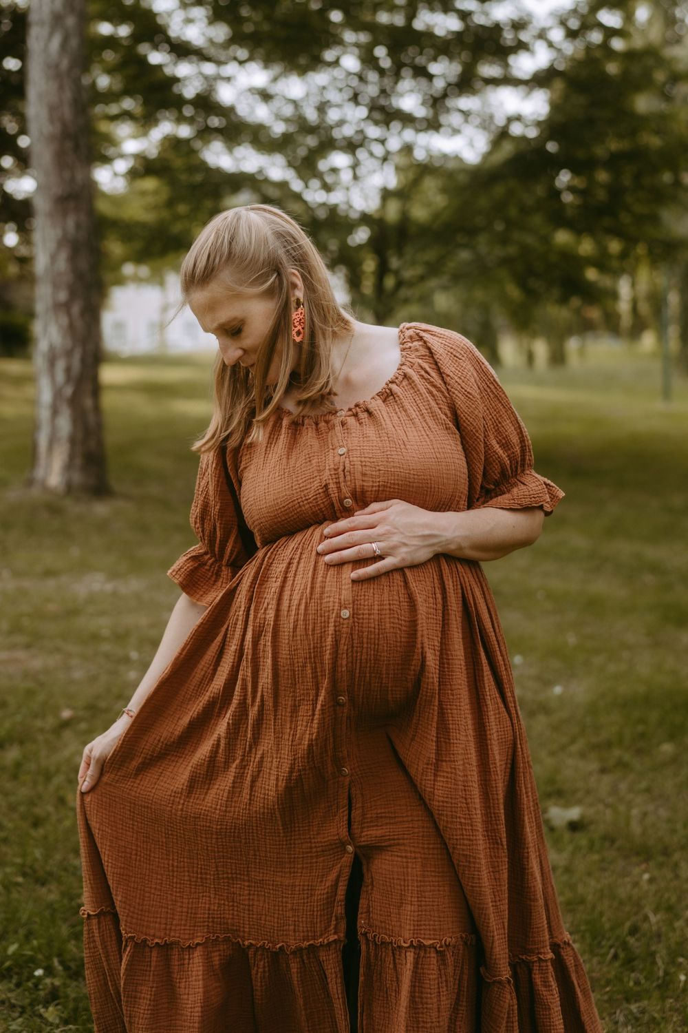 Pregnant woman in rust orange dress cradling baby bump outdoors in park.