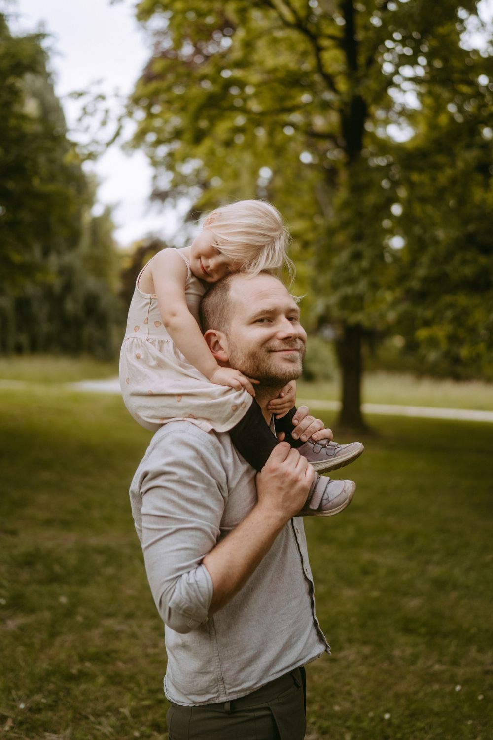 Smiling father carries blonde toddler on shoulders in lush green park.