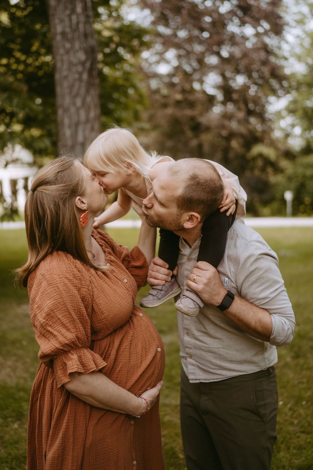 Pregnant woman in rust dress and man kiss toddler on shoulders outdoors in lush green park setting.