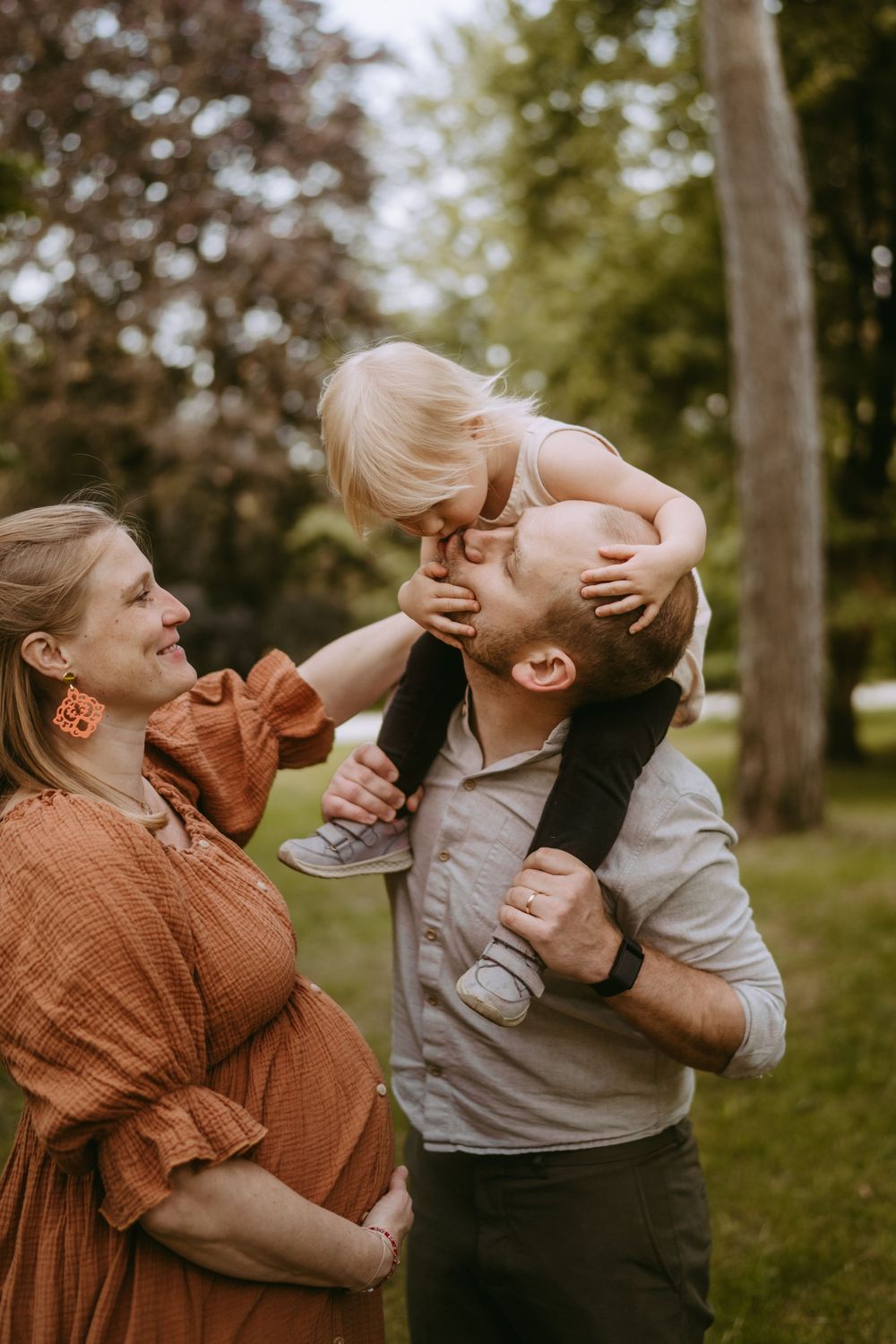 Pregnant woman smiles as man holds toddler on shoulders for a kiss in green park.