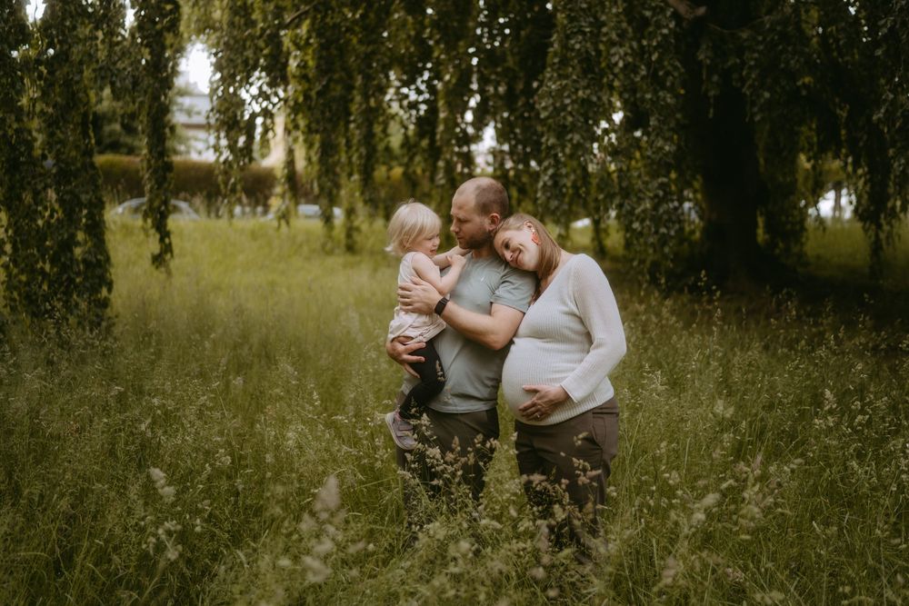 Pregnant woman and man holding toddler embrace in lush green field under weeping willow trees.