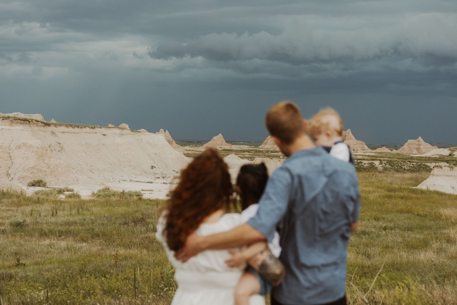 Family of four stands together gazing at dramatic stormy skies over rugged Badlands landscape.