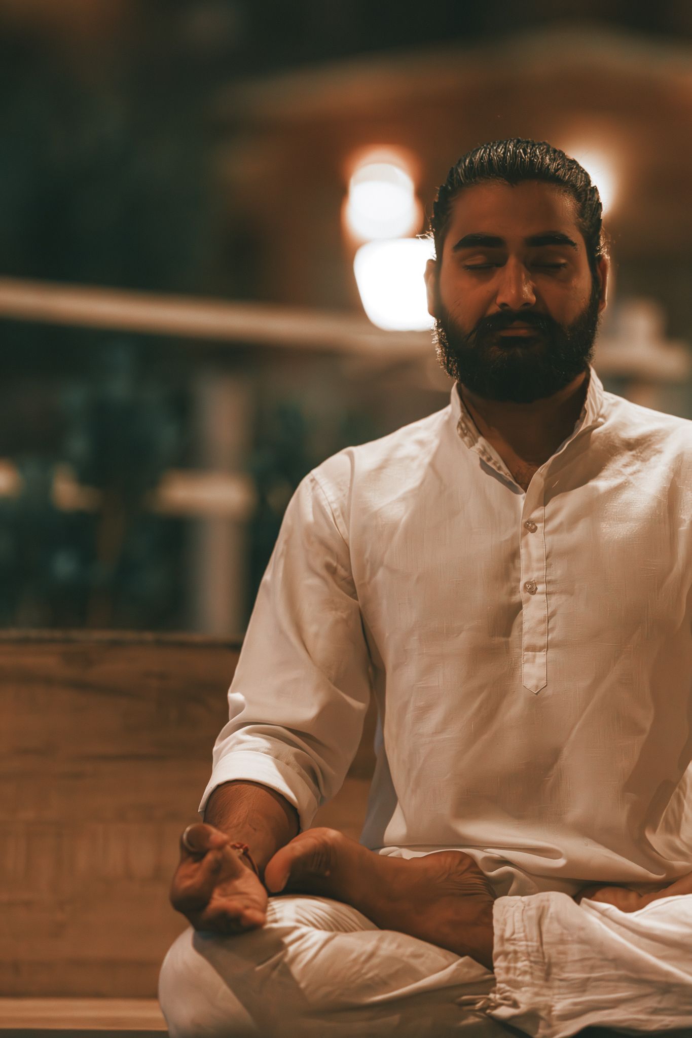Bearded man in white kurta meditates in lotus pose with eyes closed in a softly lit indoor setting.