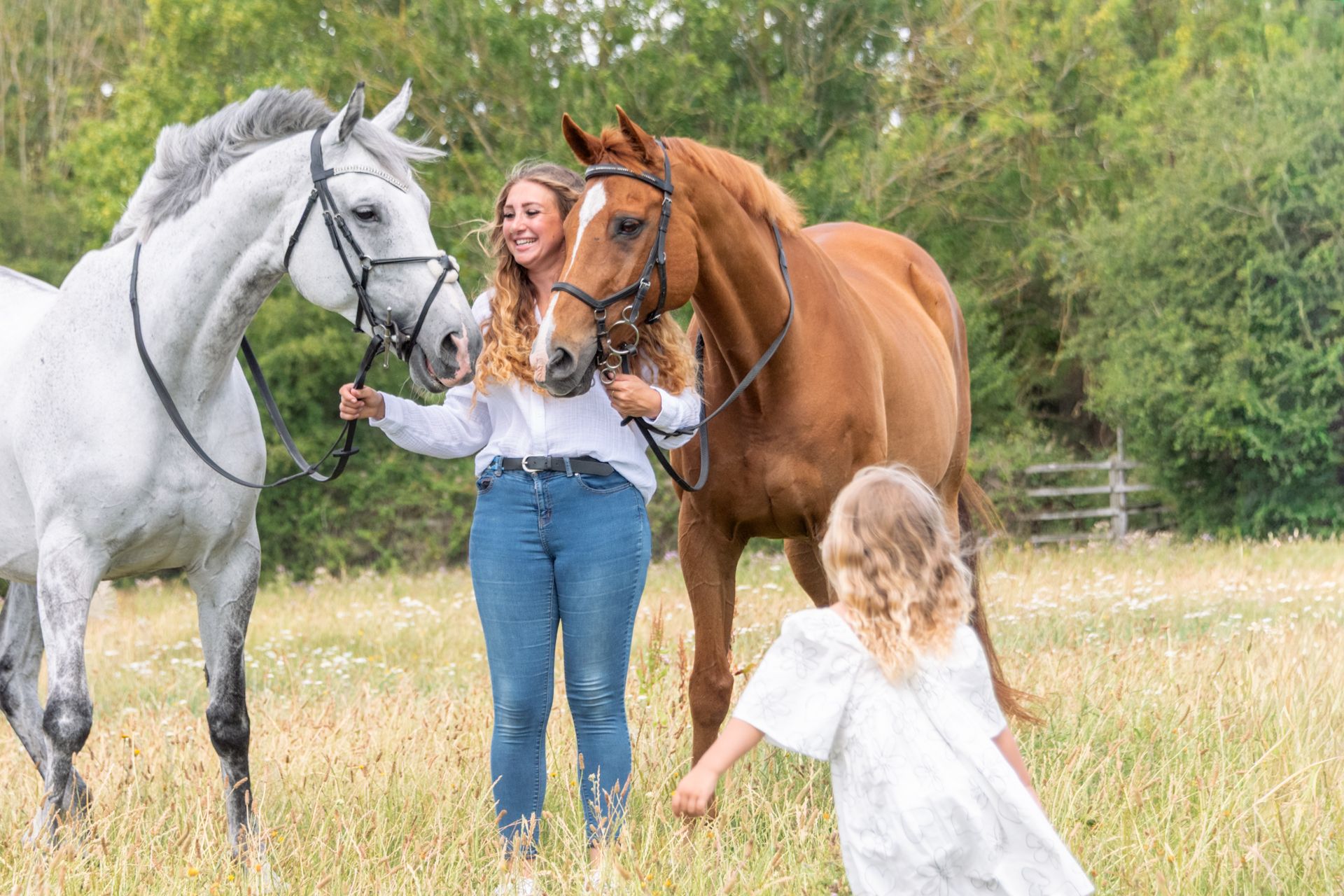 Young blonde woman in navy polo shirt standing between two grey horses wearing black bridles outdoors.