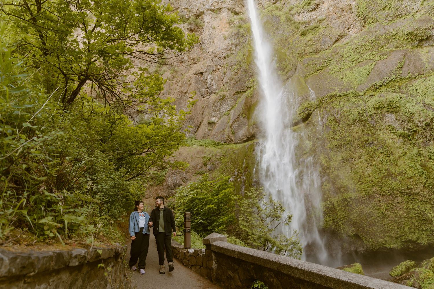 Two people stand on a stone path before a tall waterfall cascading down a moss-covered cliff surrounded by lush green trees.