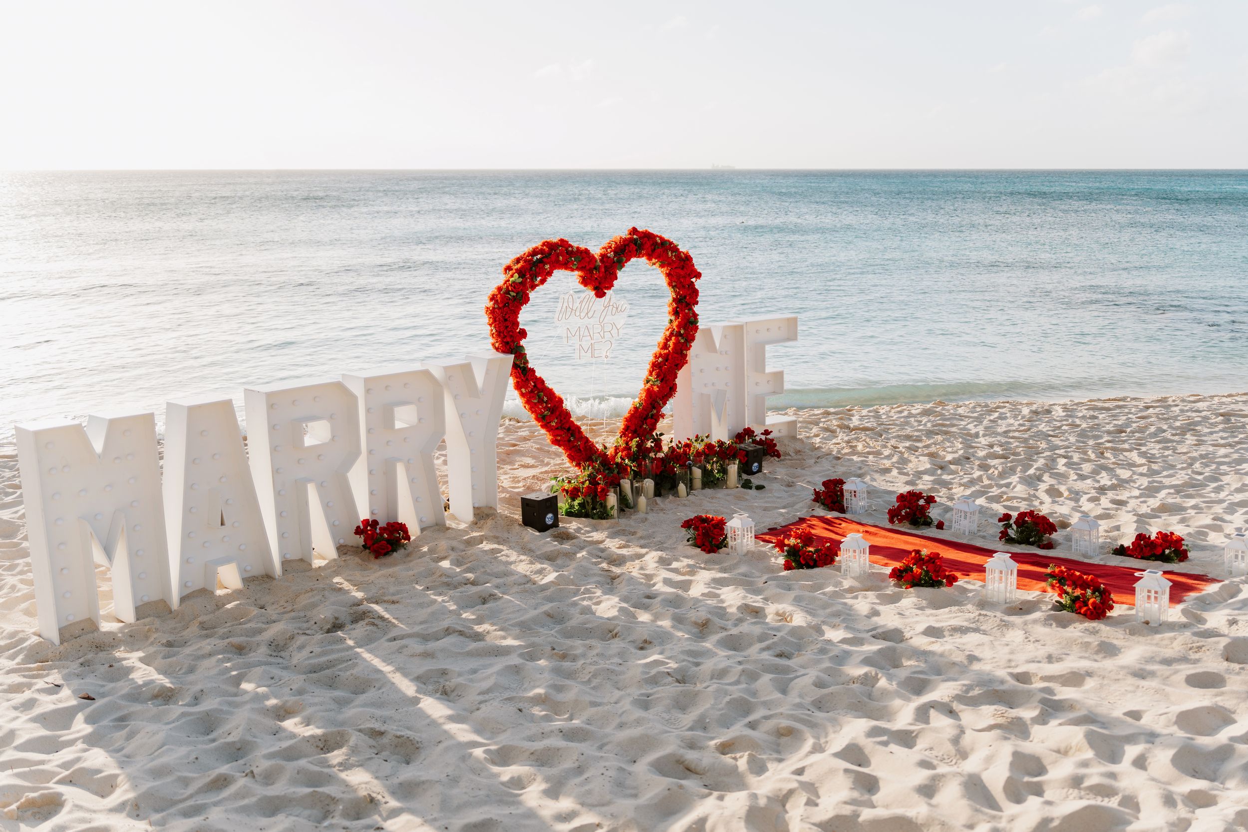 Romantic beach proposal setup with red rose heart arch, MARRY ME letters, and red floral arrangements on white sand.