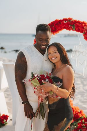 Happy couple posing on a beach with red roses, man in white outfit and woman in black dress holding a bouquet.