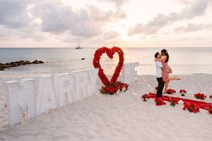 Couple kissing at romantic beach marriage proposal with red rose heart and large MARRY letters at sunset.