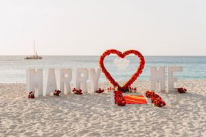 Beach marriage proposal setup with large white MARRY ME letters and a red rose heart arch on white sand by the ocean.