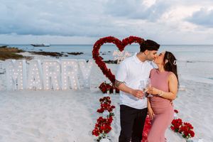 Couple sharing a romantic beach proposal with MARRY letters, red rose heart backdrop, and champagne glasses at sunset.