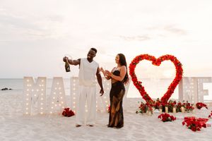 Couple celebrating beach proposal with champagne beside lit MARRY ME letters and red heart floral arch at sunset.