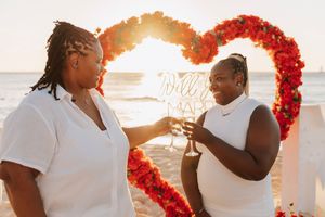 Two women in white dresses toast champagne at a beach proposal with a red rose heart arch and sunset backdrop.