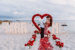 Couple kissing on beach with lit MARRY ME sign and red rose heart arch during romantic seaside proposal.