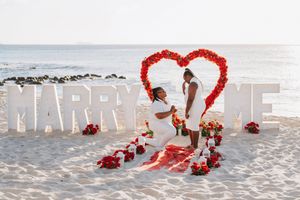 Couple sharing a marriage proposal moment on beach with red rose heart arch and MARRY ME letter decorations.