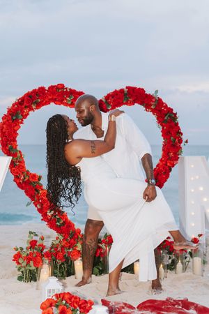 Couple embracing romantically inside a red rose heart arch on a beach with candles and floral arrangements.