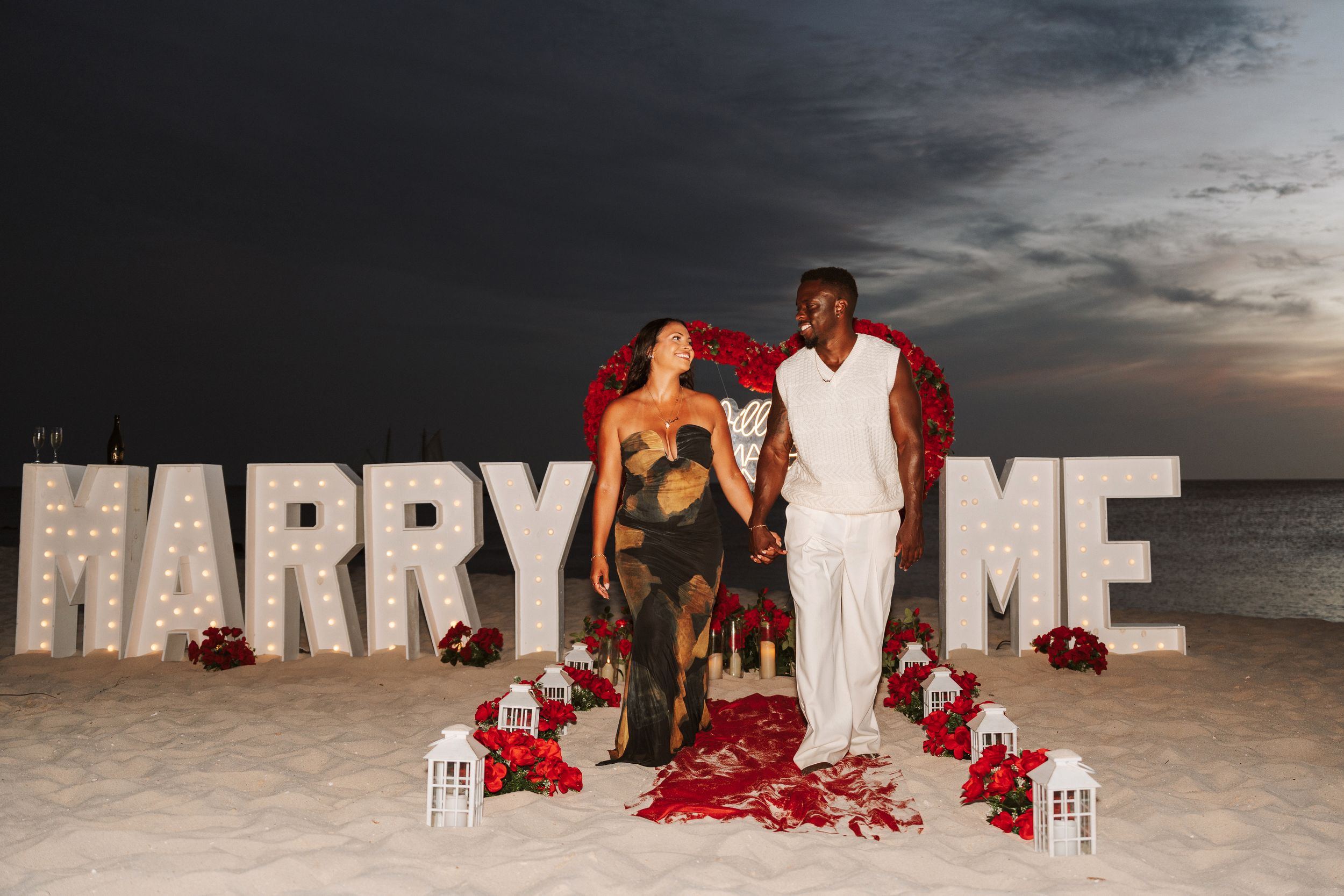 Couple stands on beach at sunset beside large illuminated MARRY ME letters surrounded by red roses and lanterns.