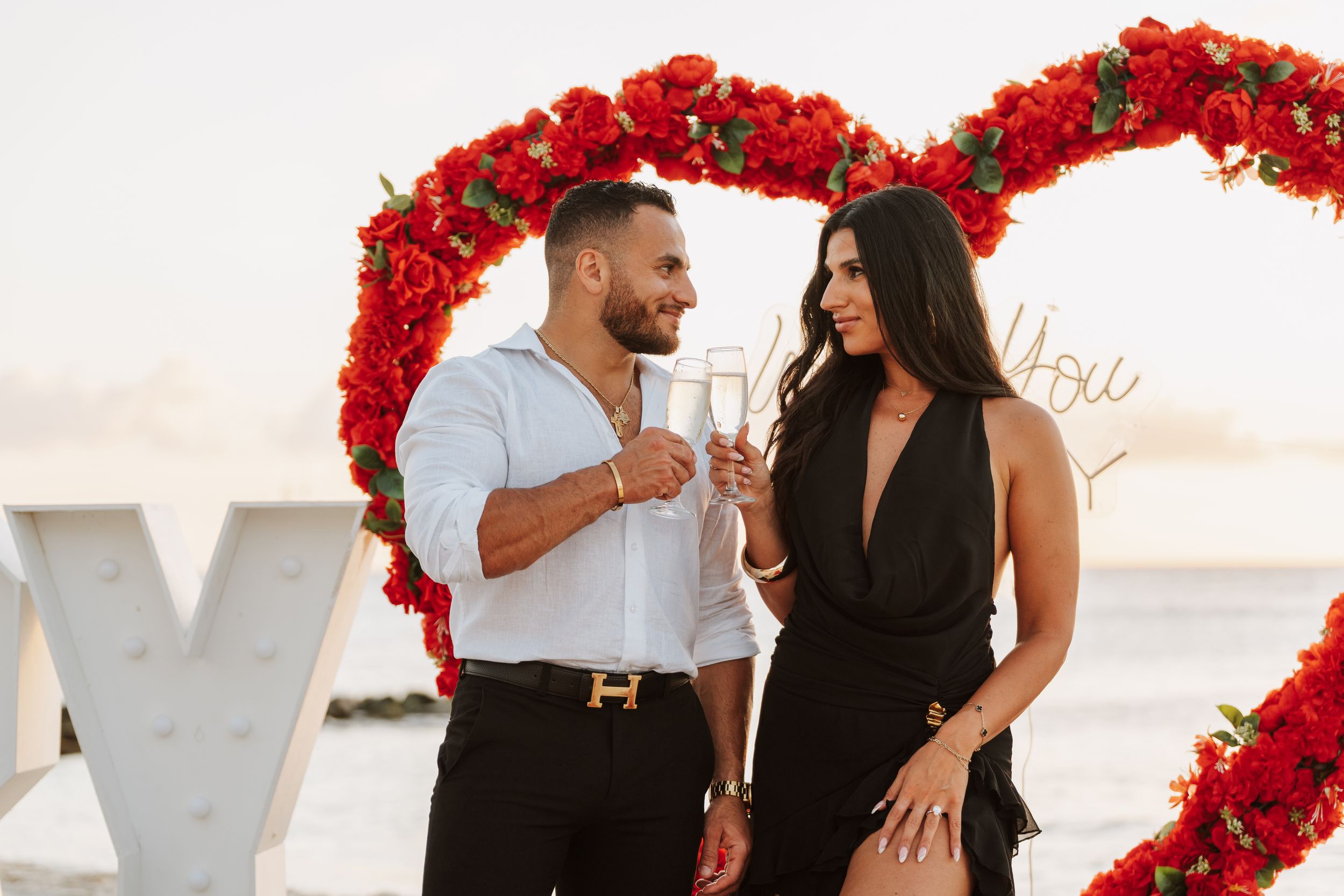 Couple toasting champagne in front of a red rose heart arch near the ocean at sunset during a romantic proposal.