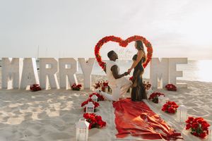 Man proposes to woman on beach with MARRY ME letters, red roses heart arch, and romantic decorations at sunset.