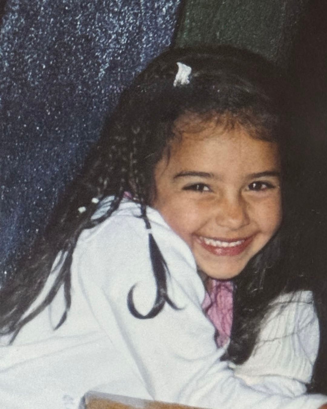 A young girl with long dark hair smiles brightly, wearing a white top with a pink shirt underneath.