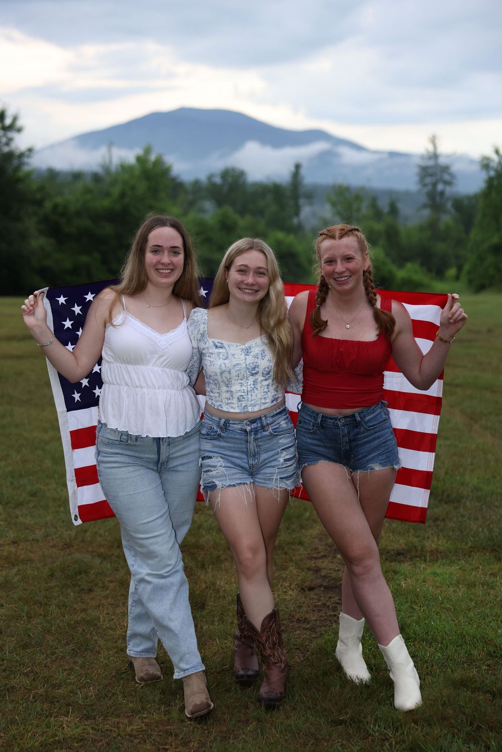Three young women holding an American flag in a green field with mountains in the background.