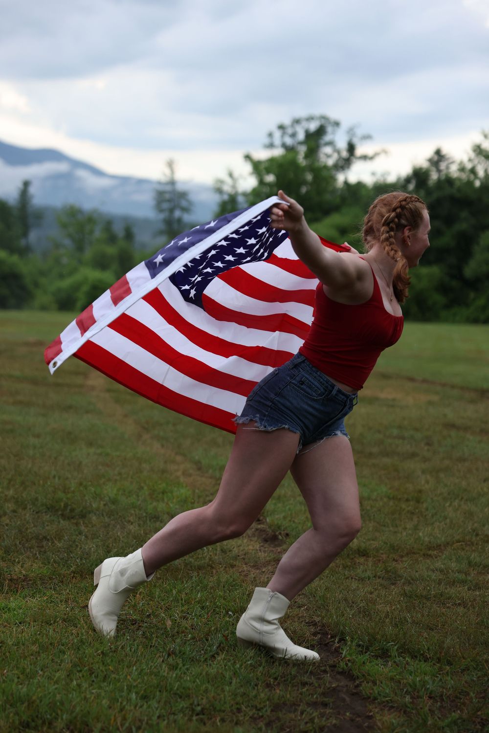 Young woman running in field holding American flag wearing red tank top and denim shorts with mountains in background.