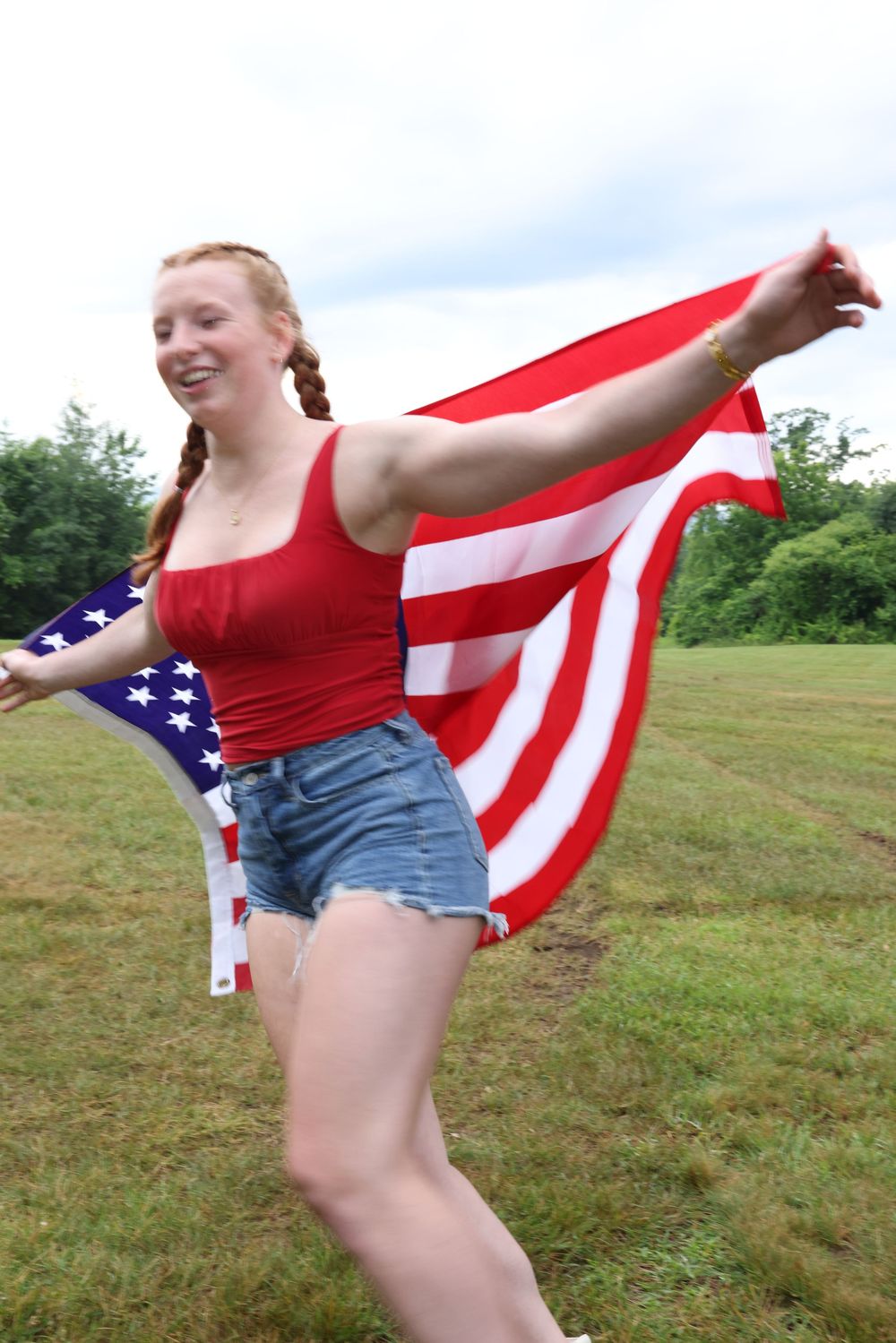 Young redhead girl running in a field holding an American flag wearing red top and denim shorts.