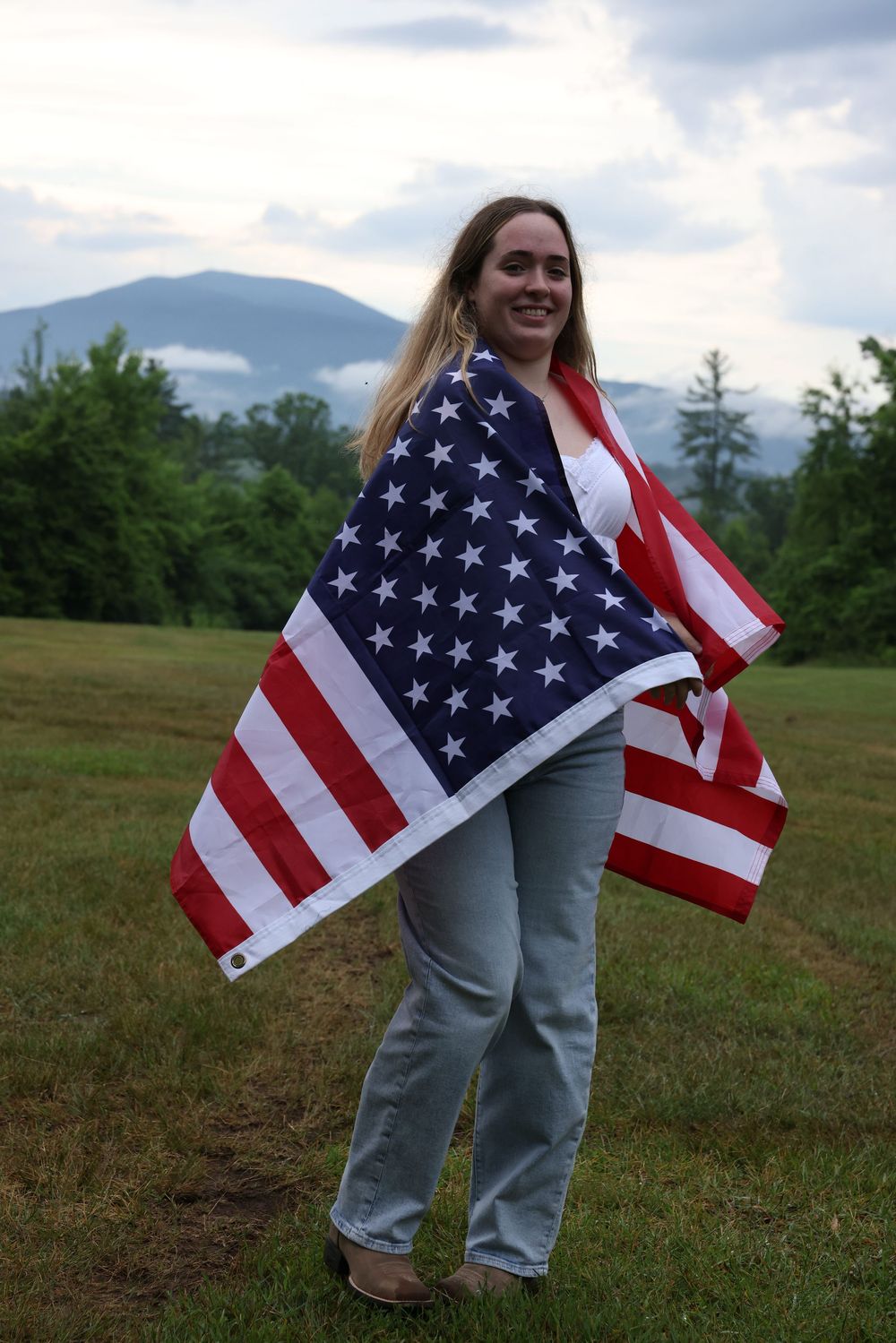 Smiling woman wrapped in American flag standing in green field with misty mountains and trees behind her.