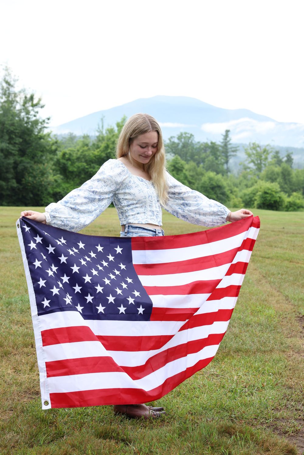 Blonde woman holding an American flag open in a green field with mountains in the background.