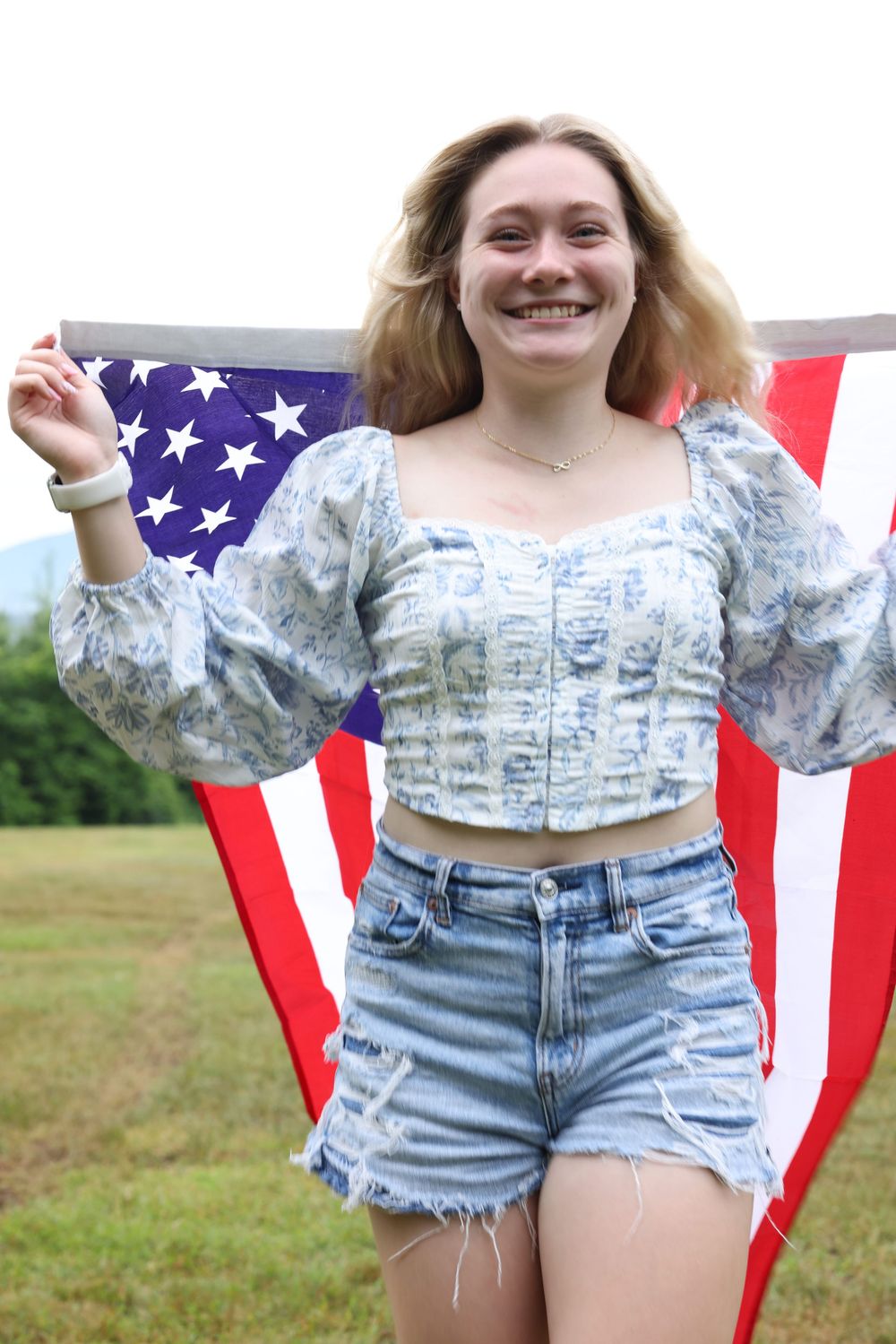 Smiling young woman holding an American flag outdoors, wearing a floral crop top and denim shorts on a green field.