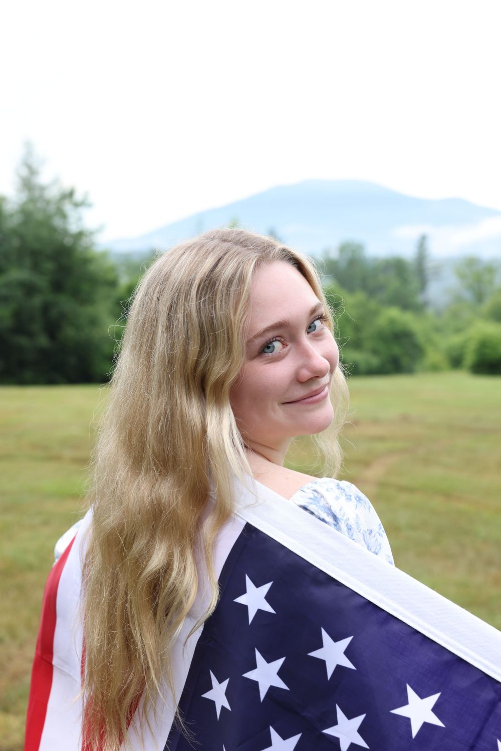 Blonde woman with long hair wrapped in American flag smiling over shoulder in outdoor field with mountain view.