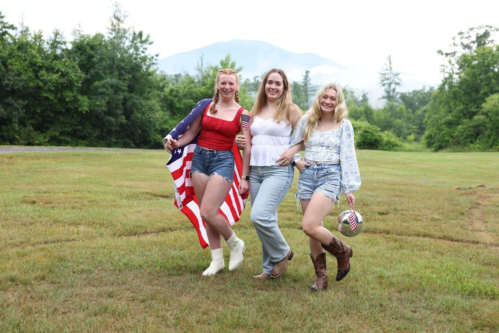 Three young women smiling outdoors holding an American flag on a green field with mountains in the background.