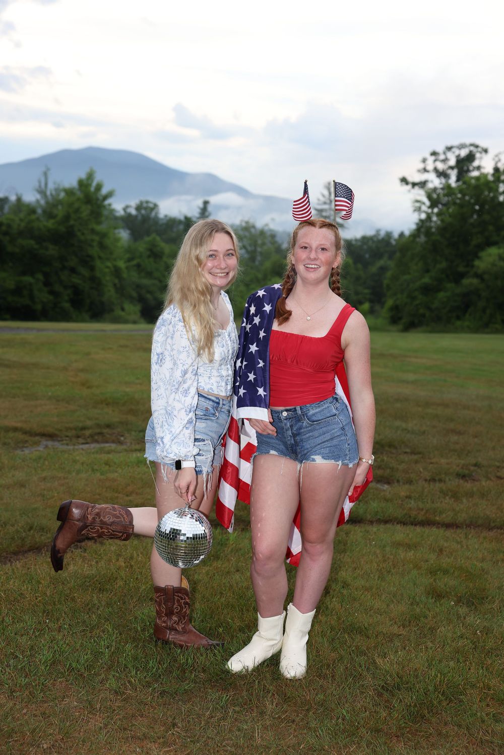 Two young women posing outdoors with American flag and disco ball, wearing patriotic outfits and cowboy boots.