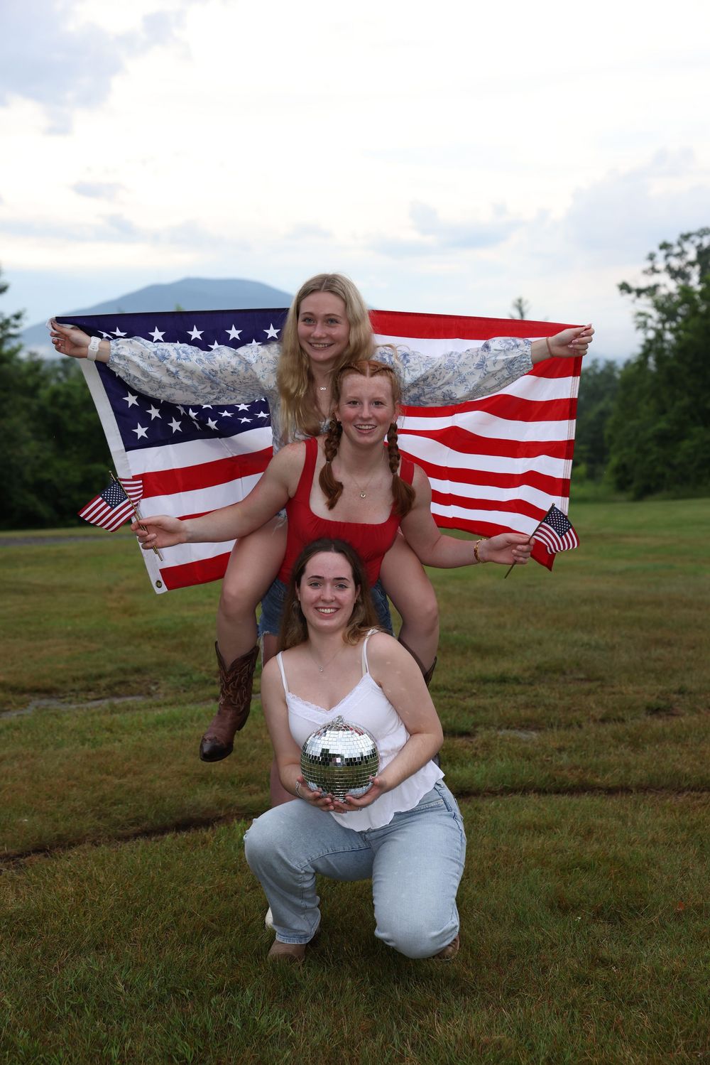 Three young women pose outdoors with American flags, one holding a mirrored ball, celebrating patriotically.