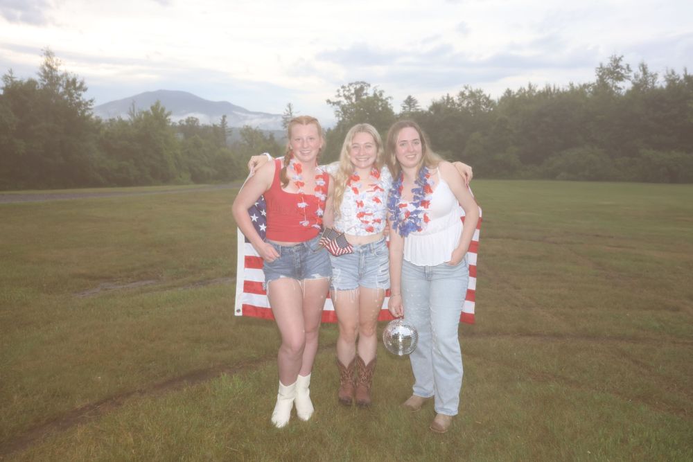 Three women pose with an American flag outdoors on a green field, dressed in patriotic red, white, and blue outfits.