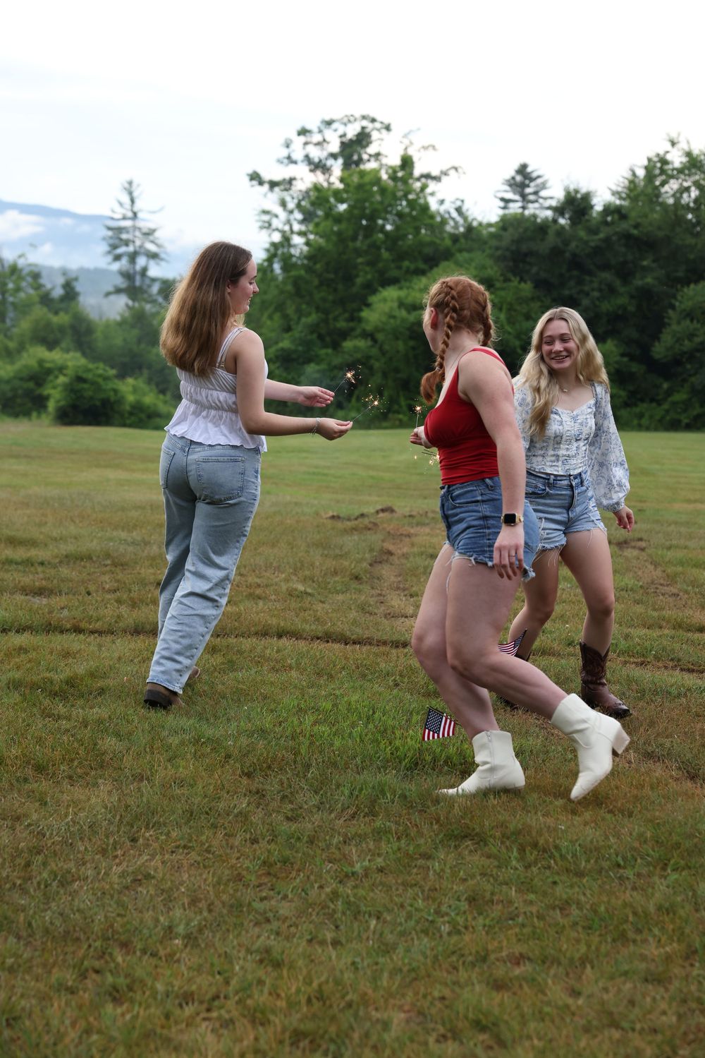 Three young women playing in a grassy field on a cloudy day near trees and mountains.