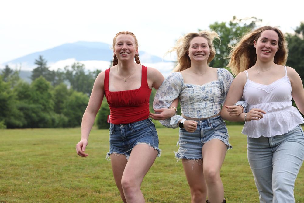 Three young women running and laughing together across a green field with mountains and trees in the background.