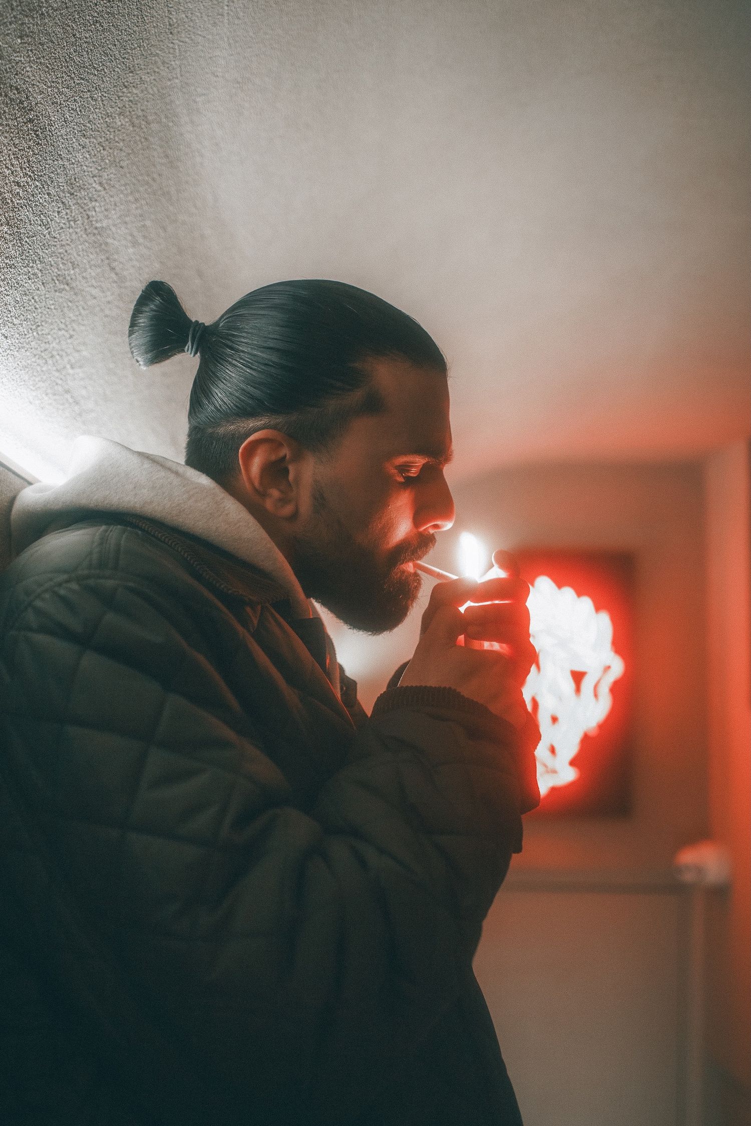 Man with hair bun lighting a cigarette in dramatic red-lit indoor setting.