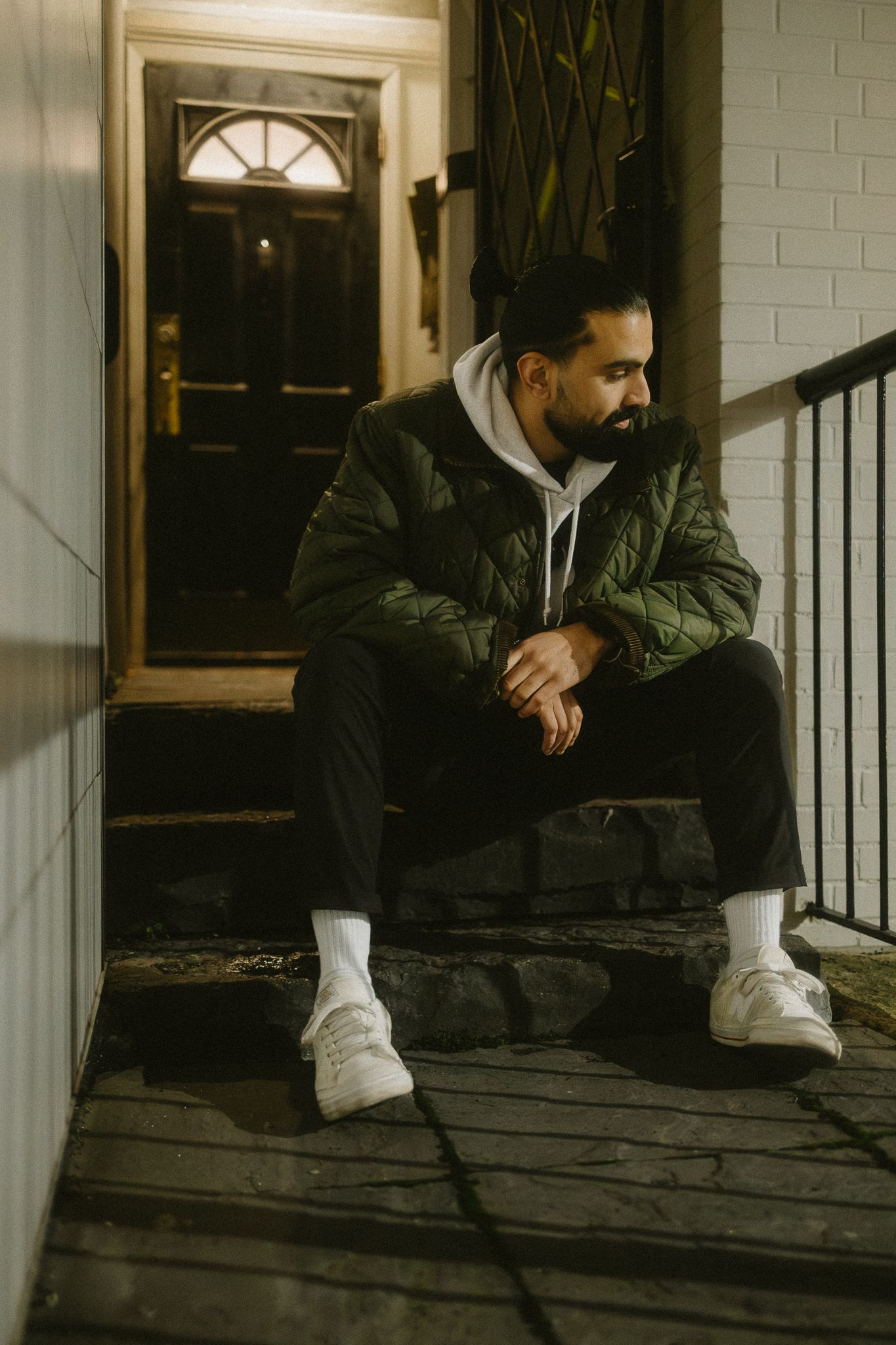 Bearded man in green quilted jacket sitting on front porch steps at night.
