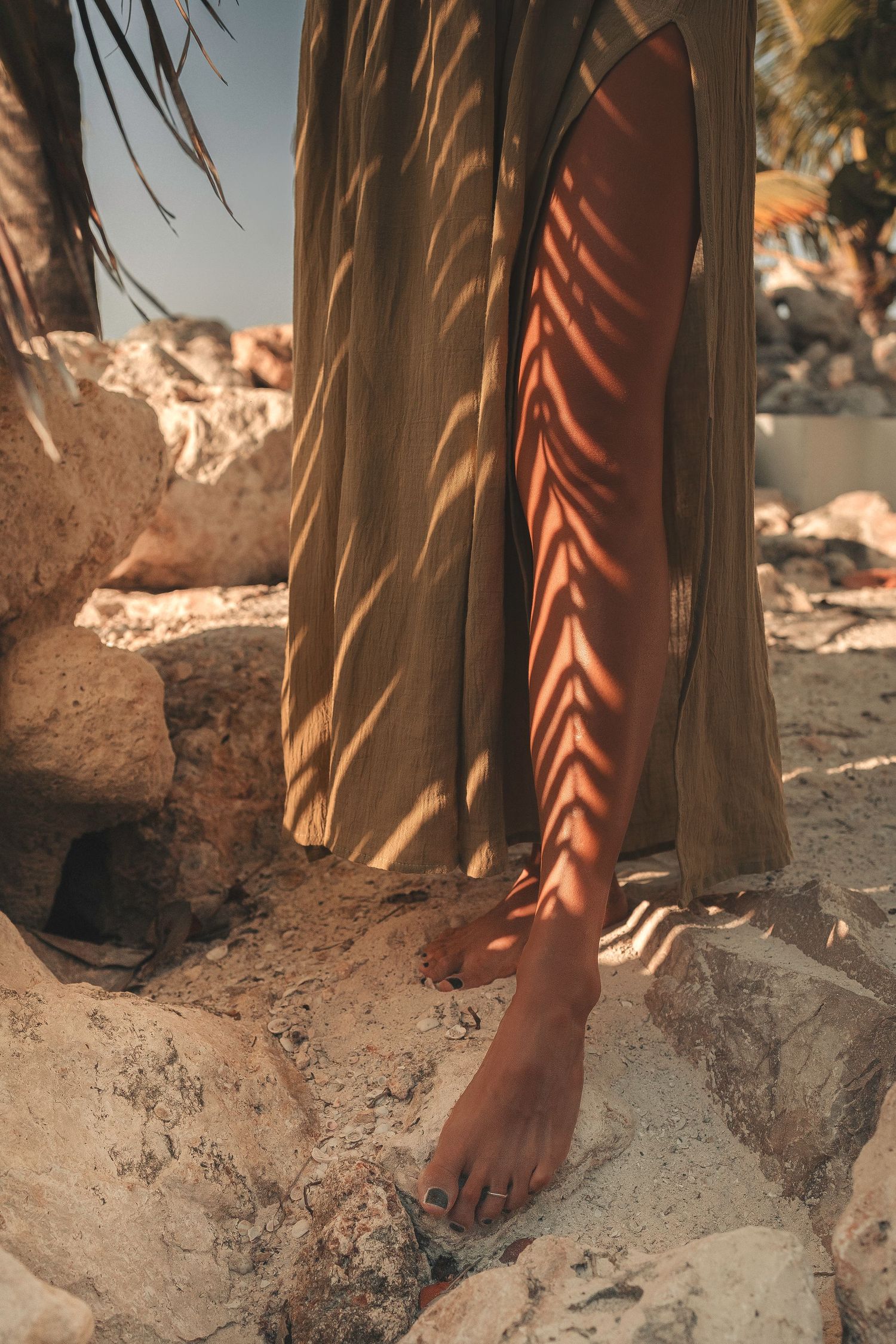 Woman in olive maxi dress with palm leaf shadows on her bare leg standing on rocks.