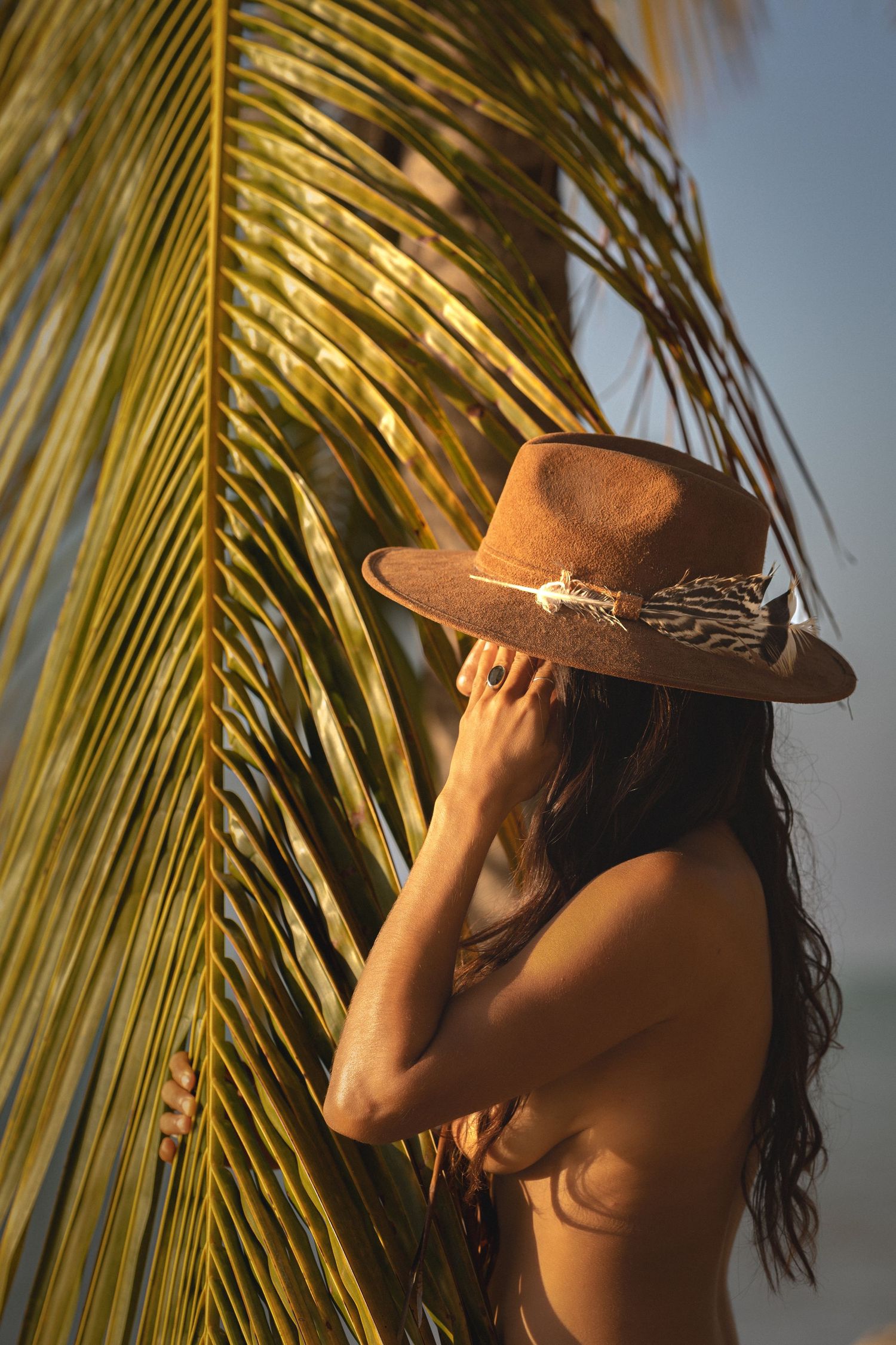Woman in brown wide-brim hat posing beside tropical palm leaf at golden sunset on beach.