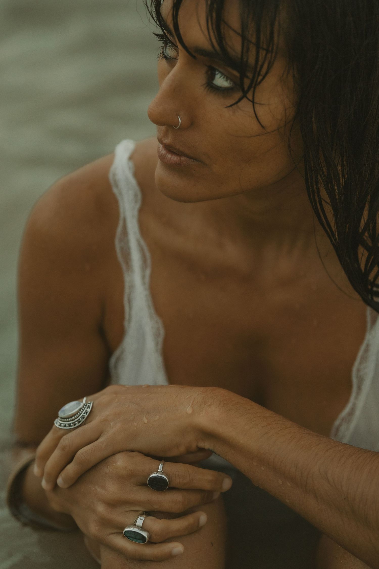 Wet-haired woman in white dress wearing silver rings, sitting in shallow ocean water.