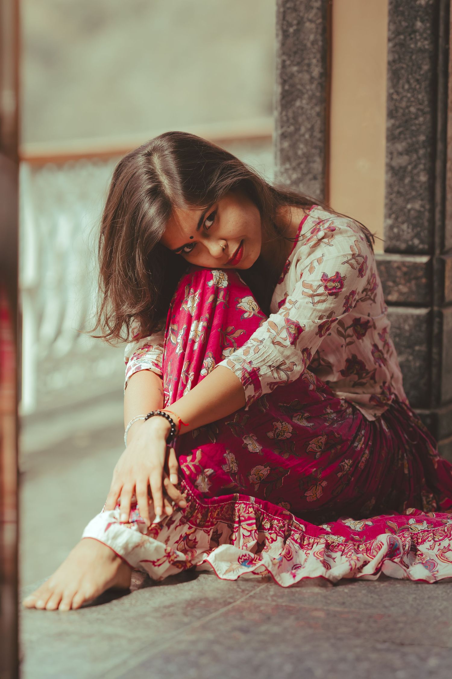 Young woman in floral pink dress sitting barefoot on floor leaning against stone pillar smiling.