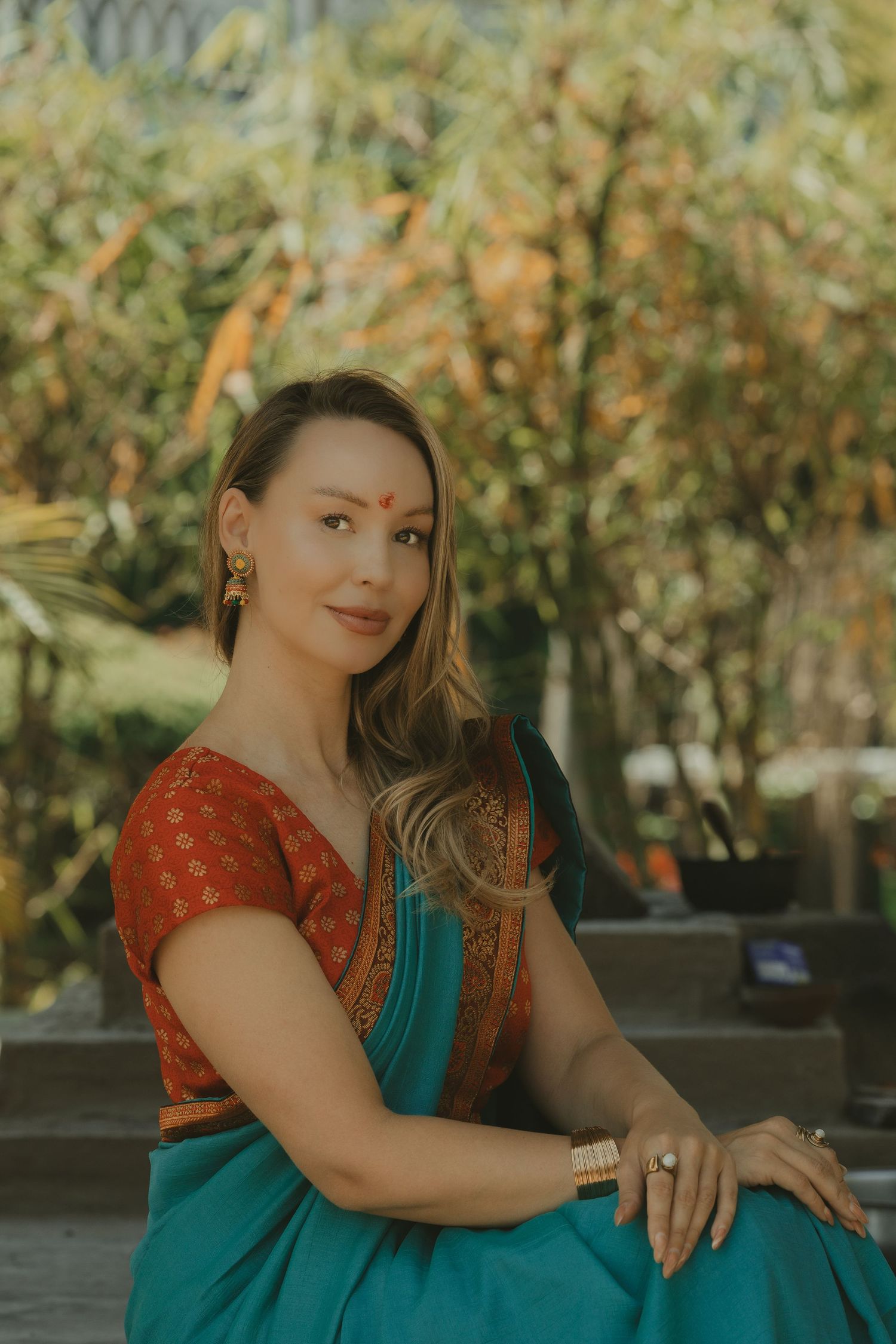 Woman wearing teal saree and red blouse with bindi sitting outdoors among autumn foliage.