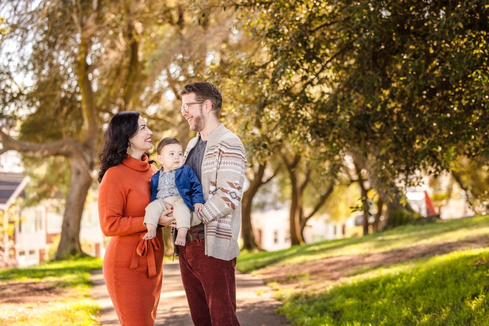 A happy young family with a baby boy poses outdoors in a sunny park surrounded by lush green trees.