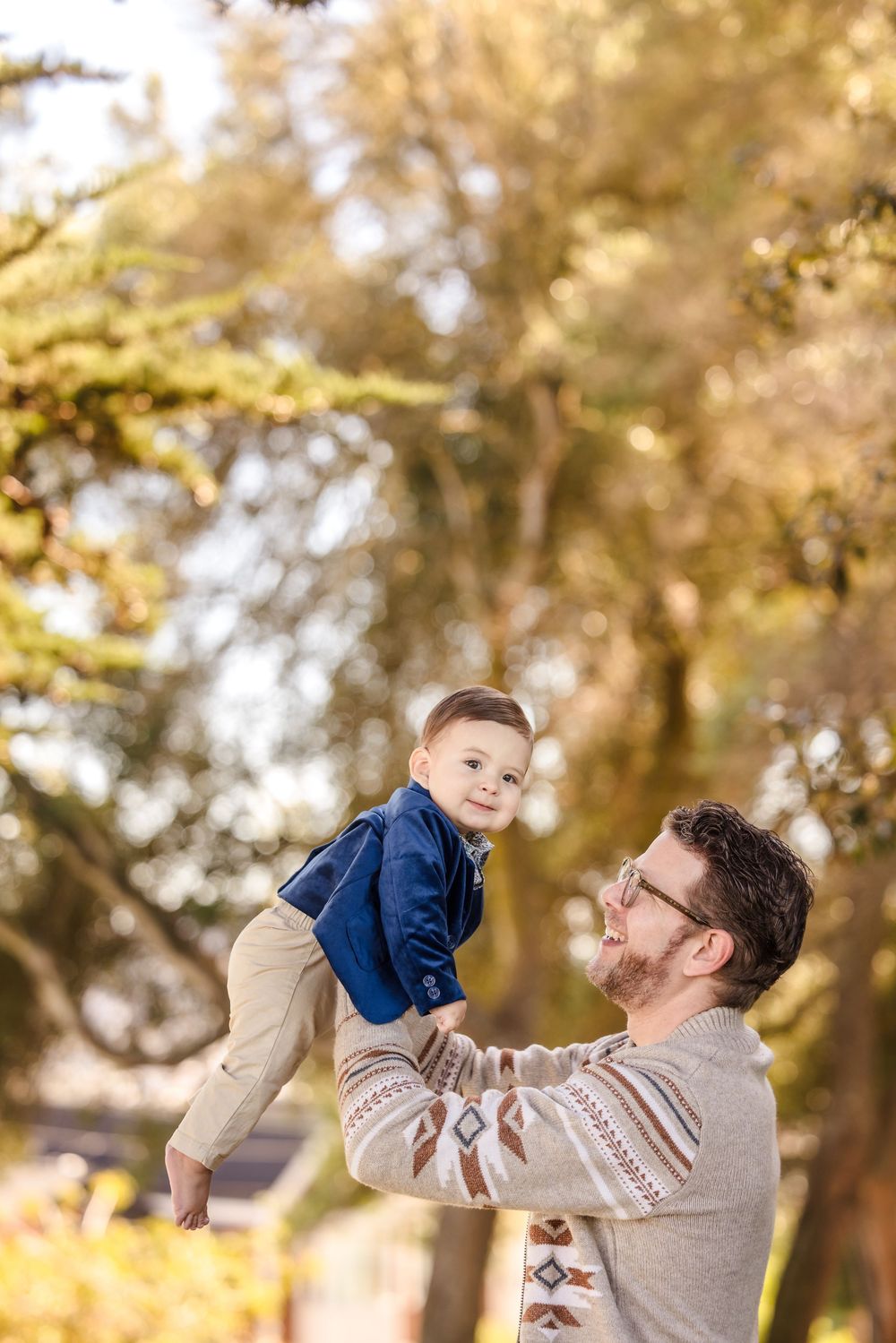 Father lifting smiling toddler son in the air outdoors among autumn trees in warm golden sunlight.