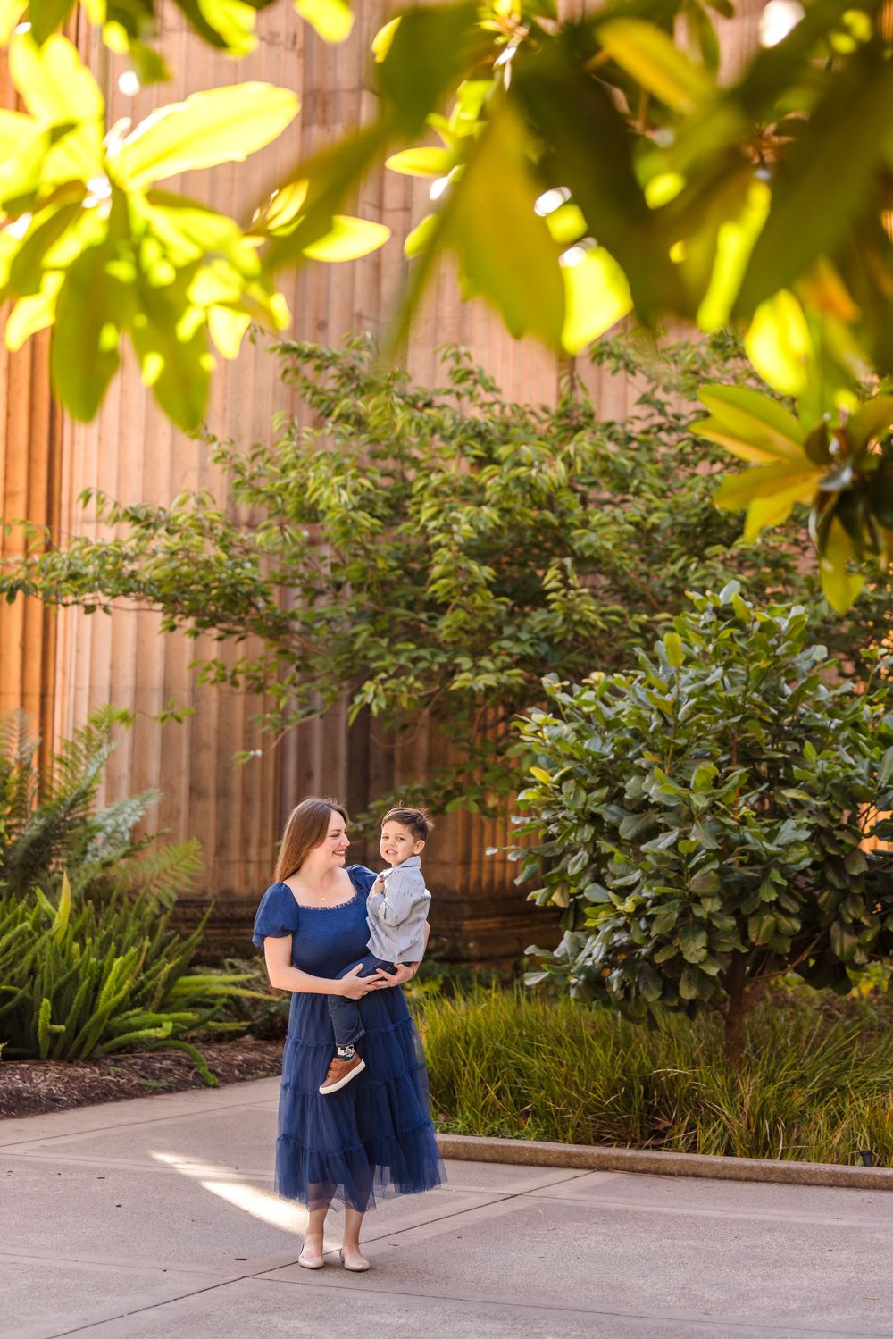 A mother in a navy blue dress holds her young son outdoors, surrounded by lush green trees and warm golden light.