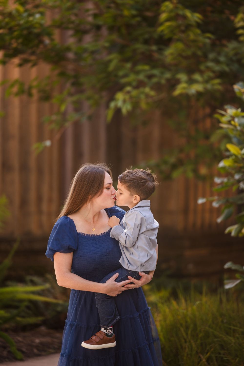 A pregnant woman in a blue dress holds her young son as they share a sweet kiss outdoors.