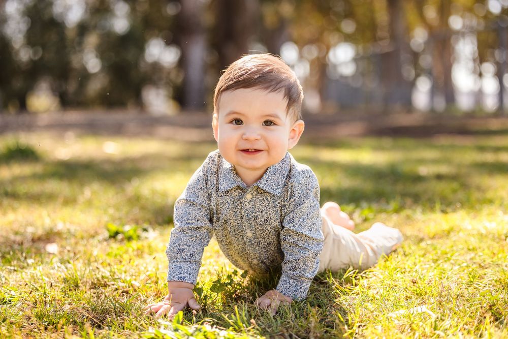 A happy baby boy crawls on green grass in a sunny park, wearing a gray sweater and beige pants.