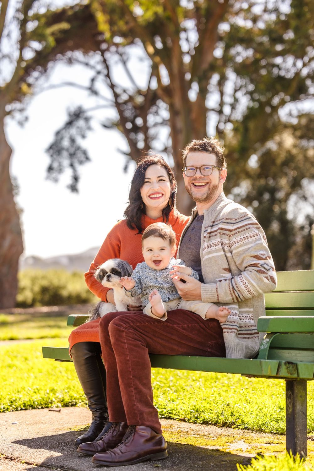 Happy family with baby and small dog sitting on green park bench outdoors on sunny day.