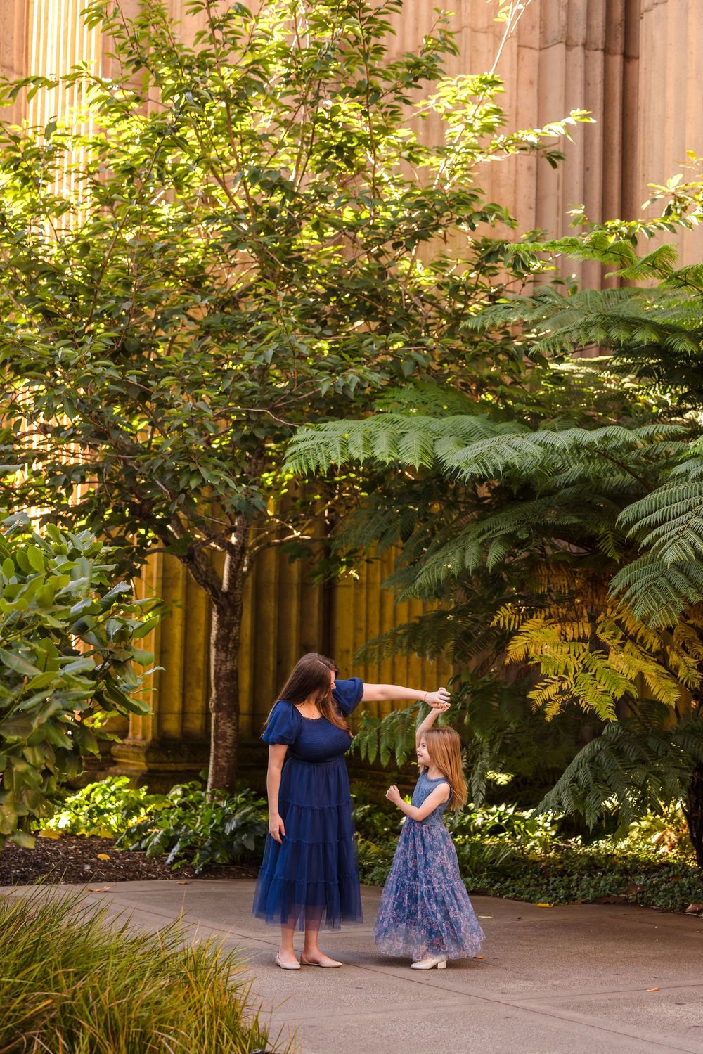 Mother in blue dress twirling her young daughter among lush greenery at the Palace of Fine Arts, San Francisco.
